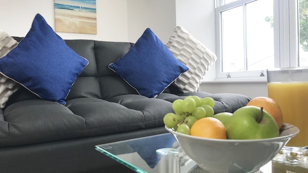 A living room with a dark grey sofa, two blue pillows, a glass coffee table, and a bowl of fruit on the table. - Lux Blu - Truro Apartment (Truro)