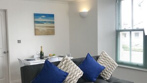 A living room with a dark sofa, two blue pillows, a white textured throw, a painting of a beach, a table with a bottle of champagne, and a window with blinds. - Lux Blu - Truro Apartment (Truro)