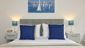 A neatly made bed with a white and blue color scheme, two bedside tables with lamps, and a painting of sailboats on the wall. - Lux Blu - Truro Apartment (Truro)