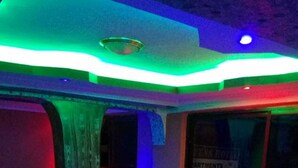 Una habitación tenuemente iluminada con piso a cuadros, un sofá de cuero negro y una zona de bar con un letrero que dice 'PARTMENTS'.