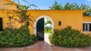 Um edifício amarelo com porta e arco preto, cercado por vegetação e flores laranjas.