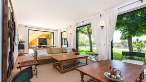 Uma sala de estar ampla com sofá, mesa de centro e mesa de jantar. Há uma televisão e uma janela com cortinas.