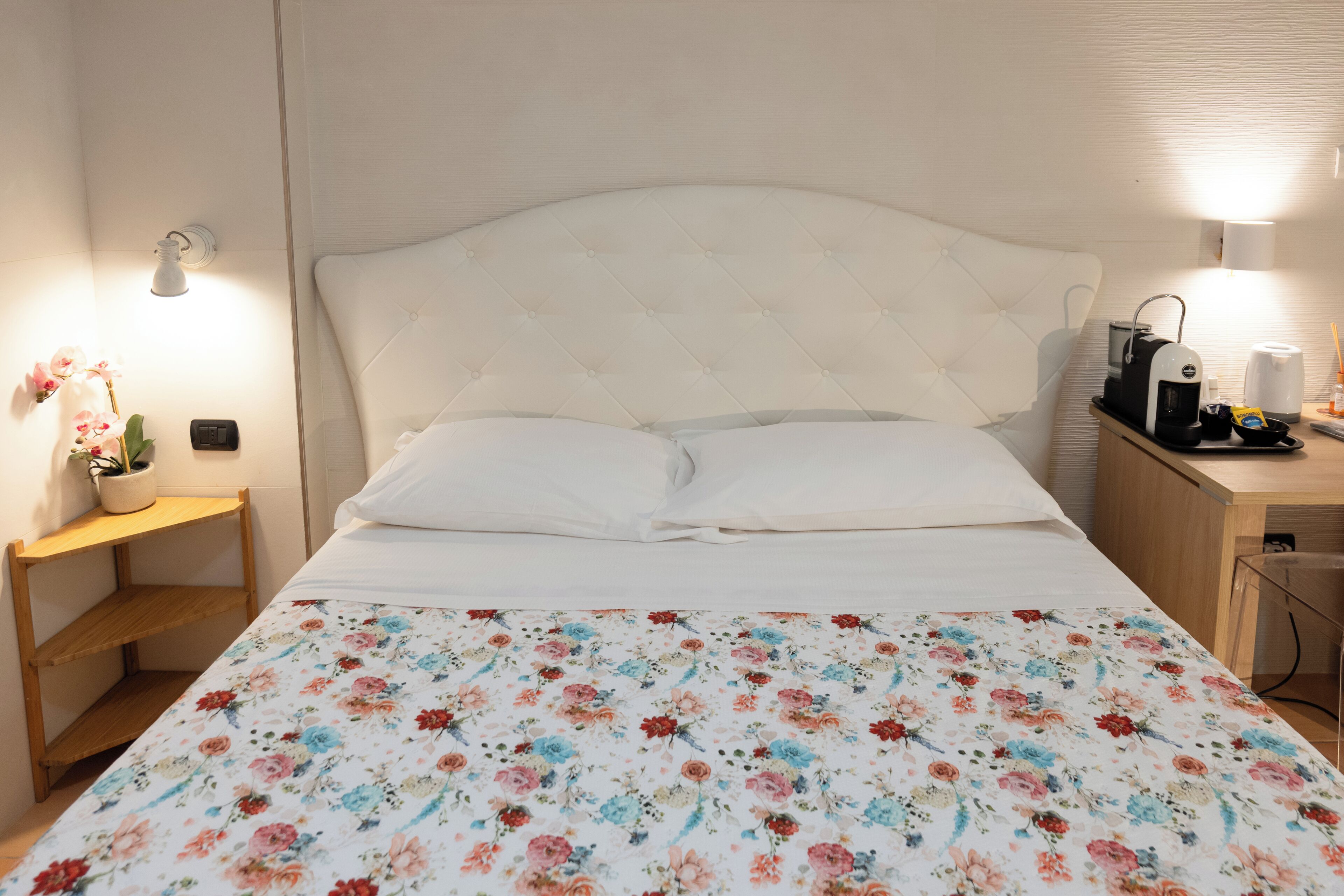 A neatly made bed with a floral bedspread, a bedside lamp, a small table with a potted plant, and a desk with a coffee maker and other items.