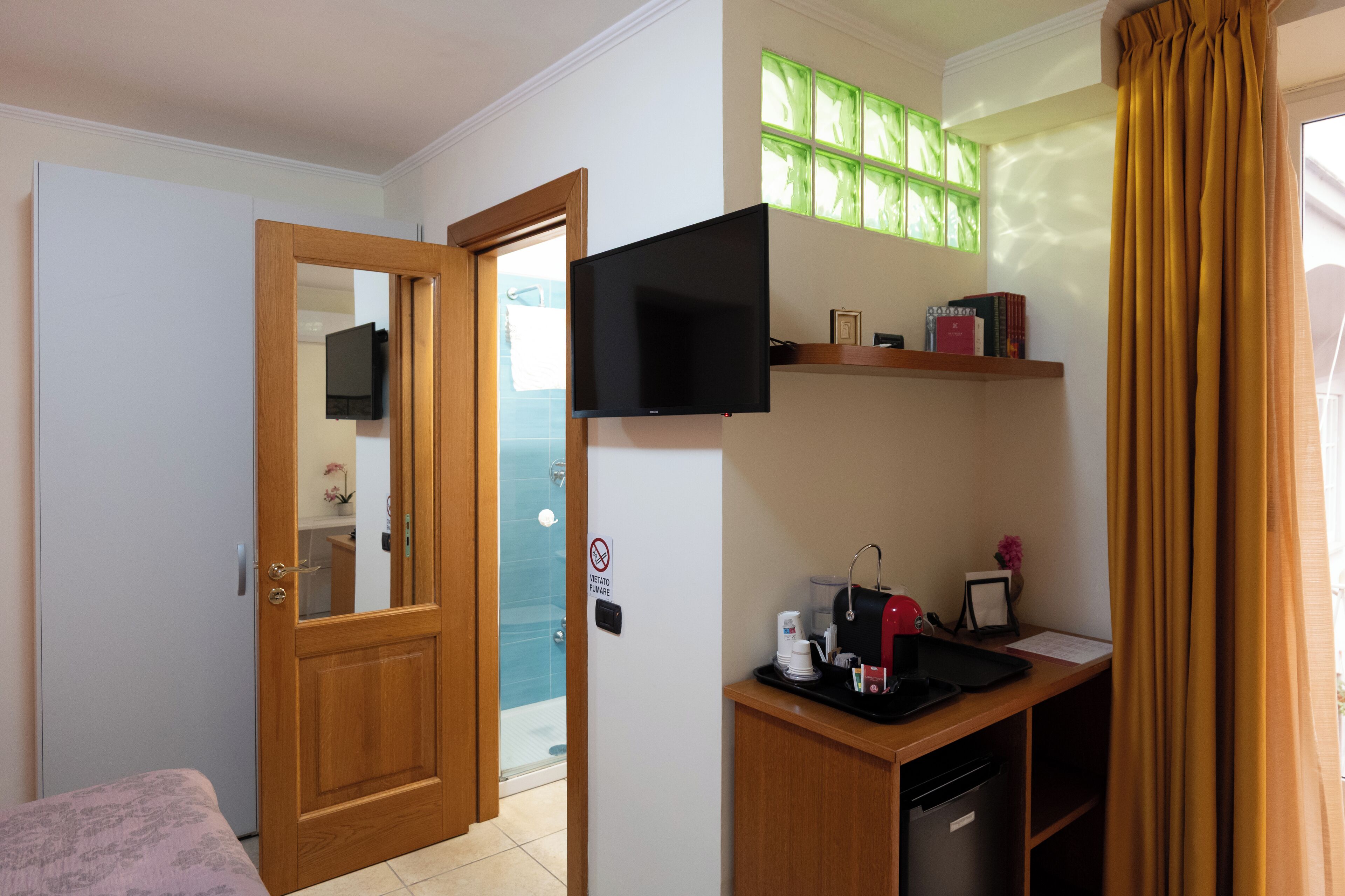 A hotel room with a wooden door, a television mounted above a desk, a shower area, and a window with green-tinted glass.