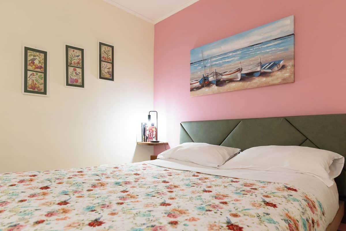 A bedroom with a floral bedspread, a green upholstered headboard, and a painting of boats on the wall.