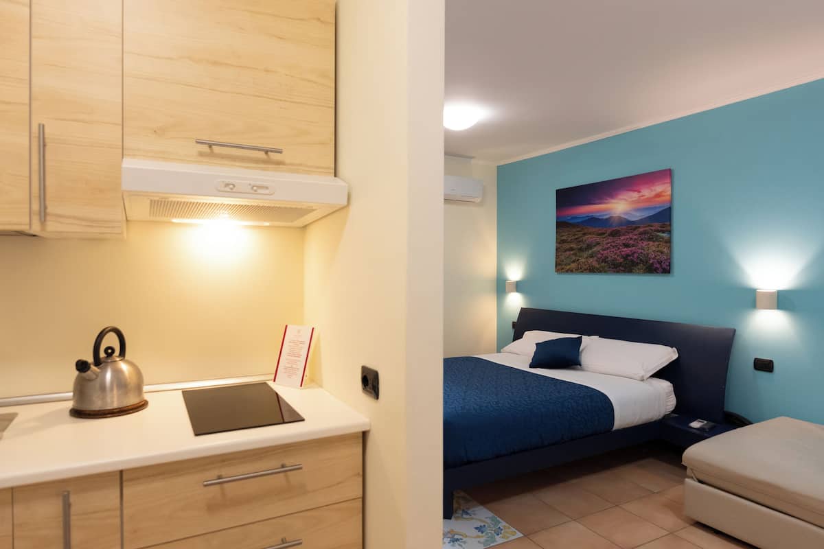 A room with a kitchenette, a bed with a blue headboard, and a painting on the wall.