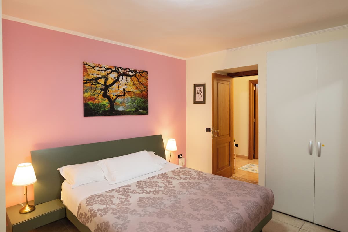 A bedroom with a bed, nightstands, a painting, a wardrobe, and a door leading to another room.