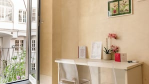 Una habitación con un escritorio blanco, dos sillas transparentes, un ventanal con vistas a edificios, una planta en maceta e ilustración botánica enmarcada en la pared.