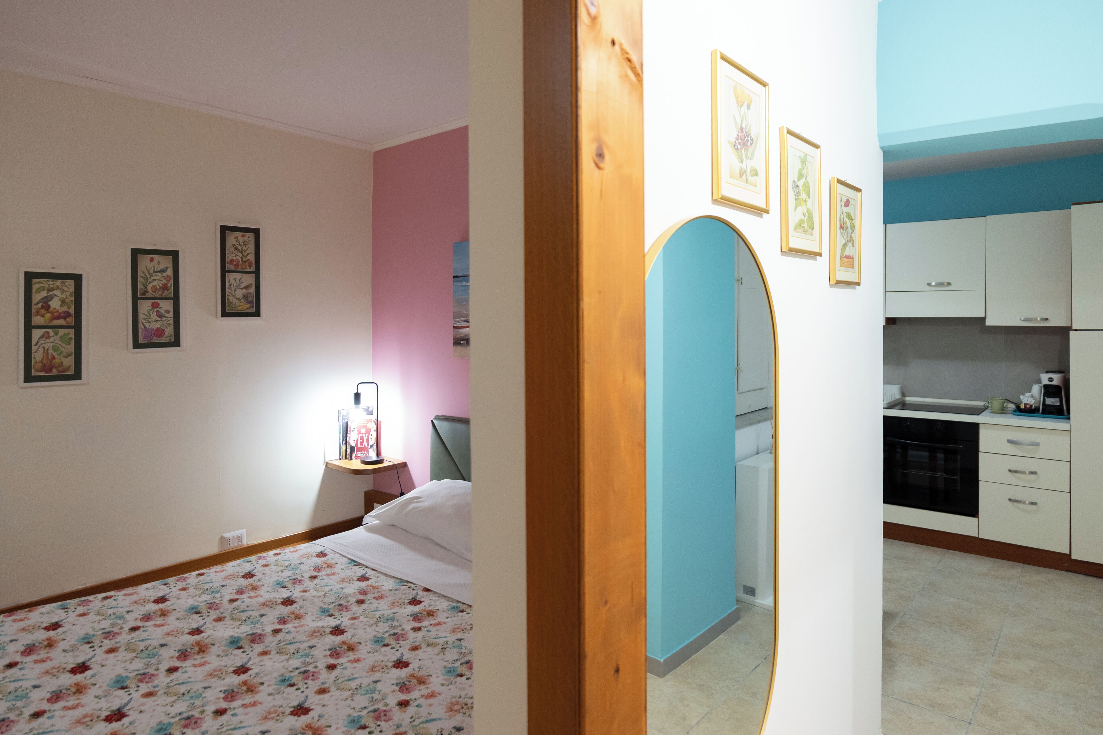 A bedroom with a floral bedspread, a bedside table with a lamp, and a pink wall. A doorway leads to a kitchen with white cabinets and a blue accent wall.