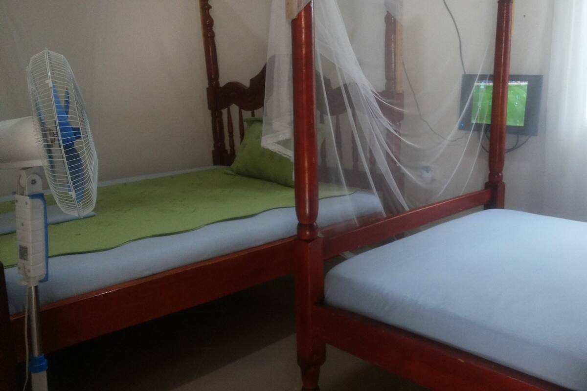A room with two single beds, a bedside table with a fan, and a TV mounted on the wall.