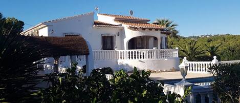 A white villa with a tiled roof, a balcony, and a view of a lush green landscape.