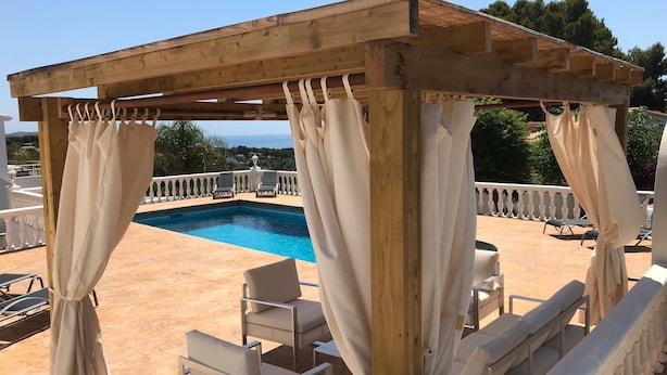 A wooden pergola with white curtains, a pool, and lounge chairs.