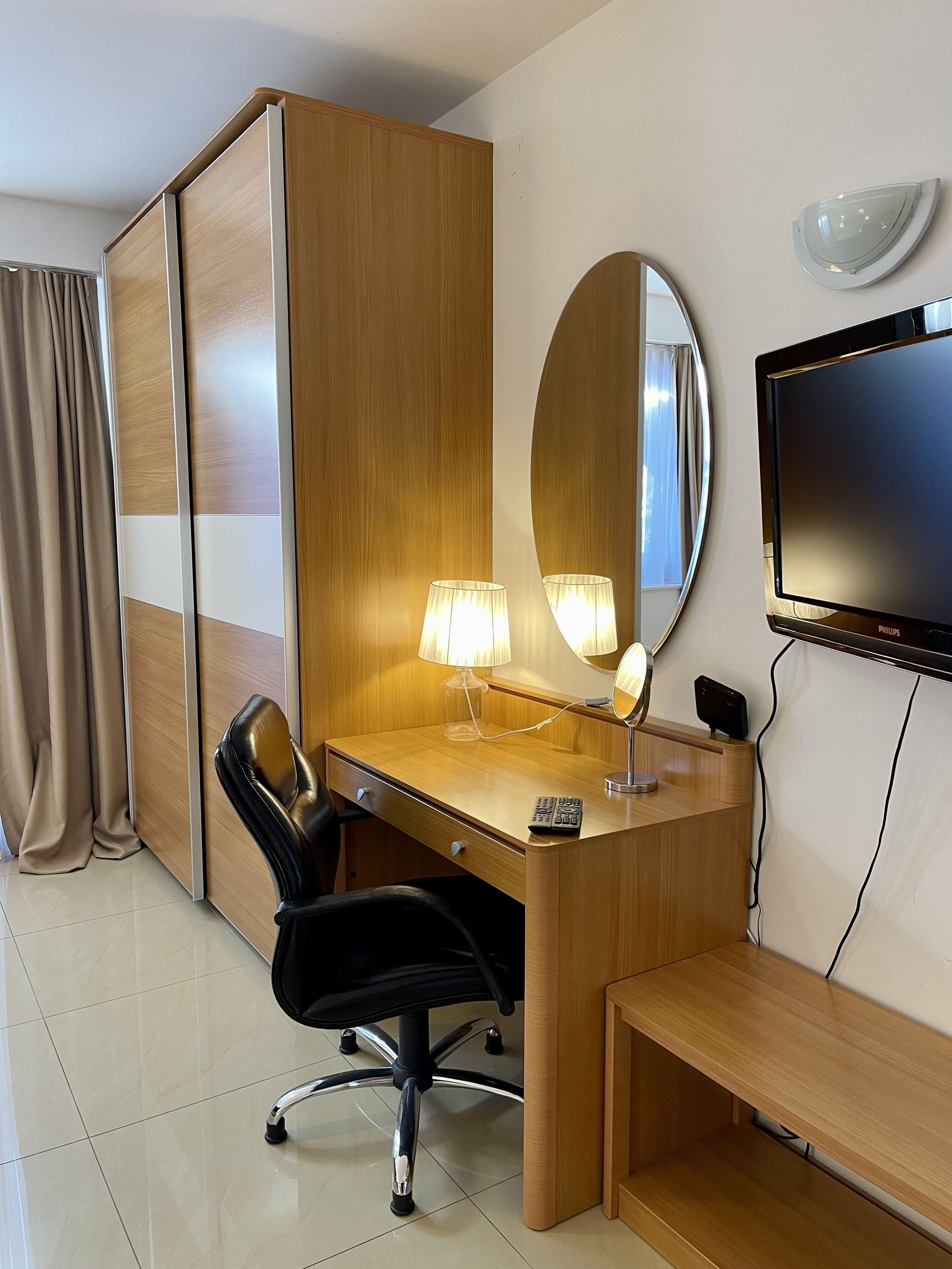 A hotel room with a wooden desk, a black office chair, a bedside table, a TV, and a mirror.