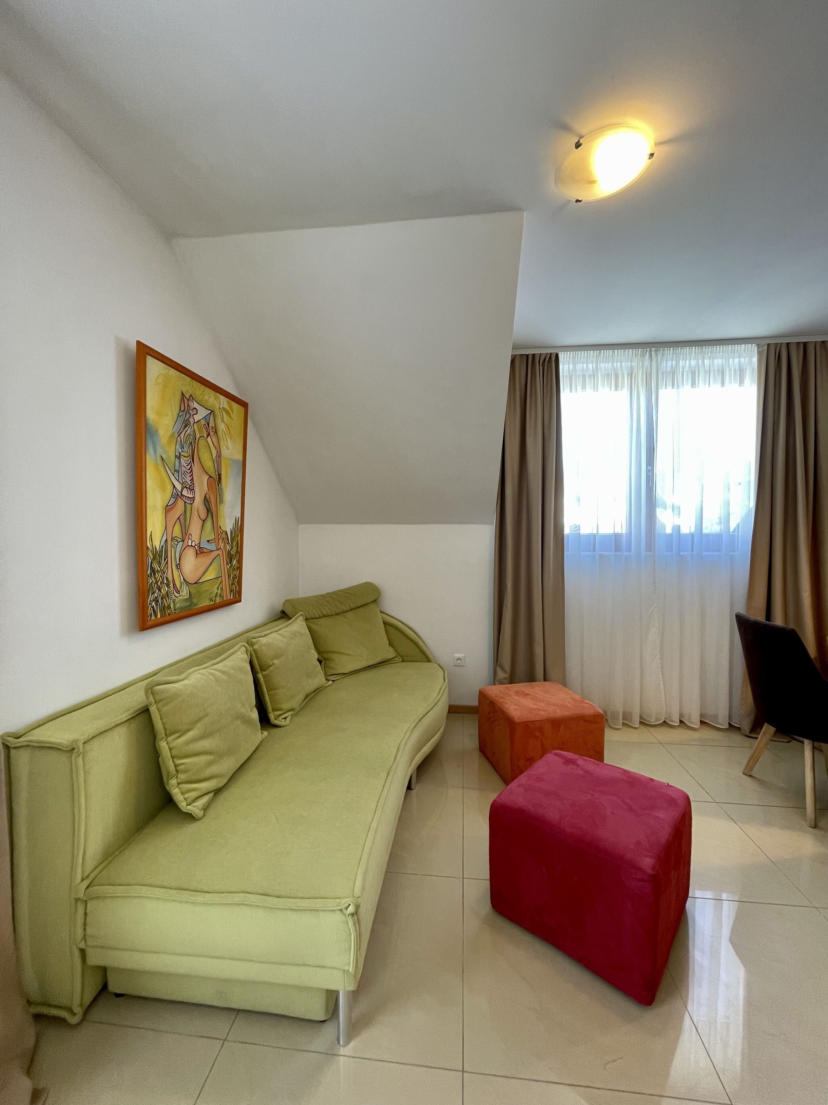 A living room with a green sofa, a red ottoman, a black chair, a painting on the wall, and a window with curtains.