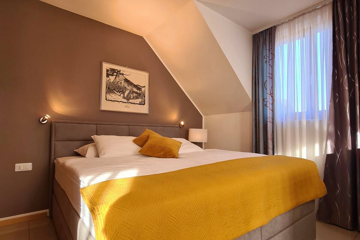 A bedroom with a large bed, a headboard, a wall-mounted light, a framed picture, and a window with curtains.