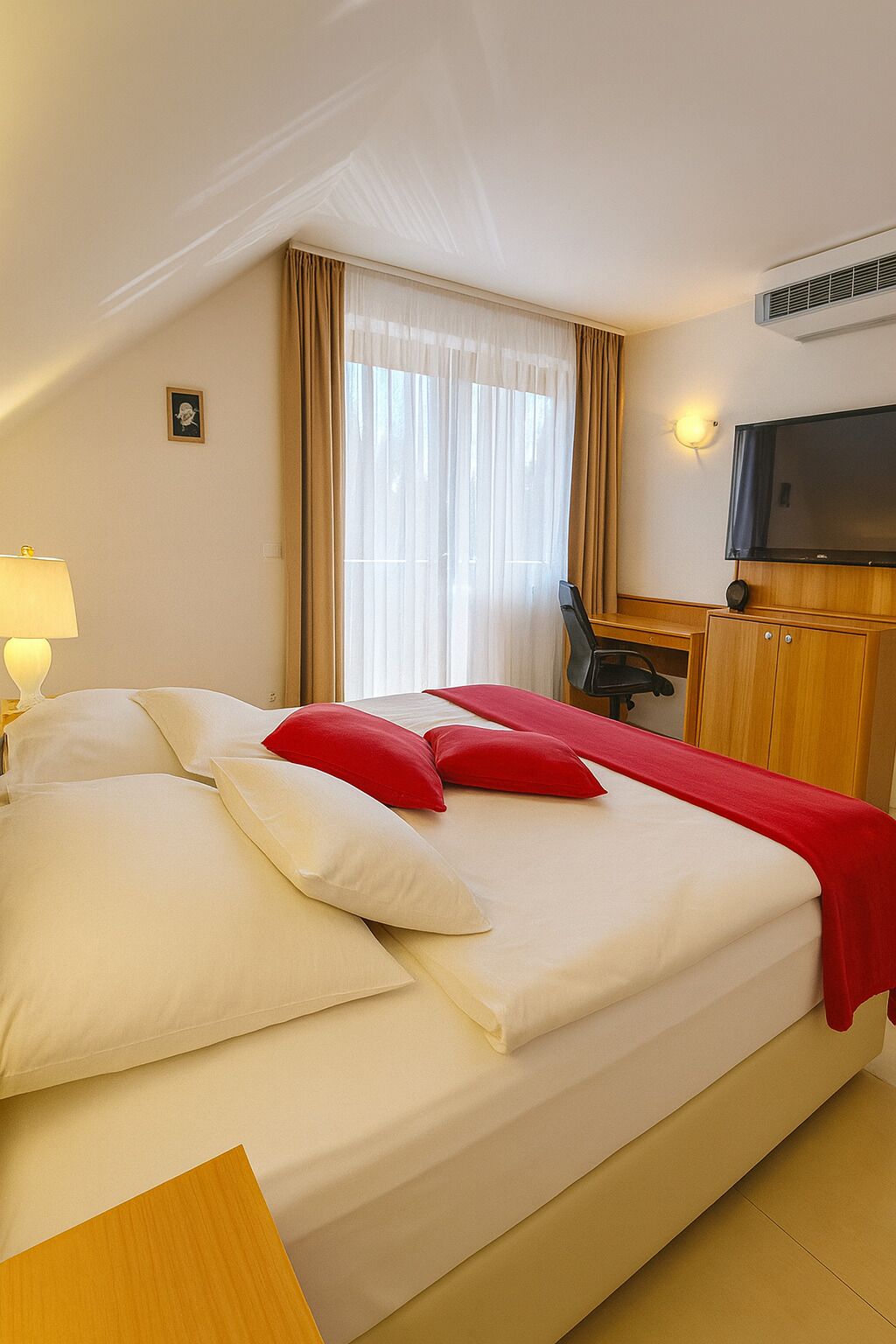 Habitación de hotel con cama, escritorio, silla, televisión y ventana con cortinas.