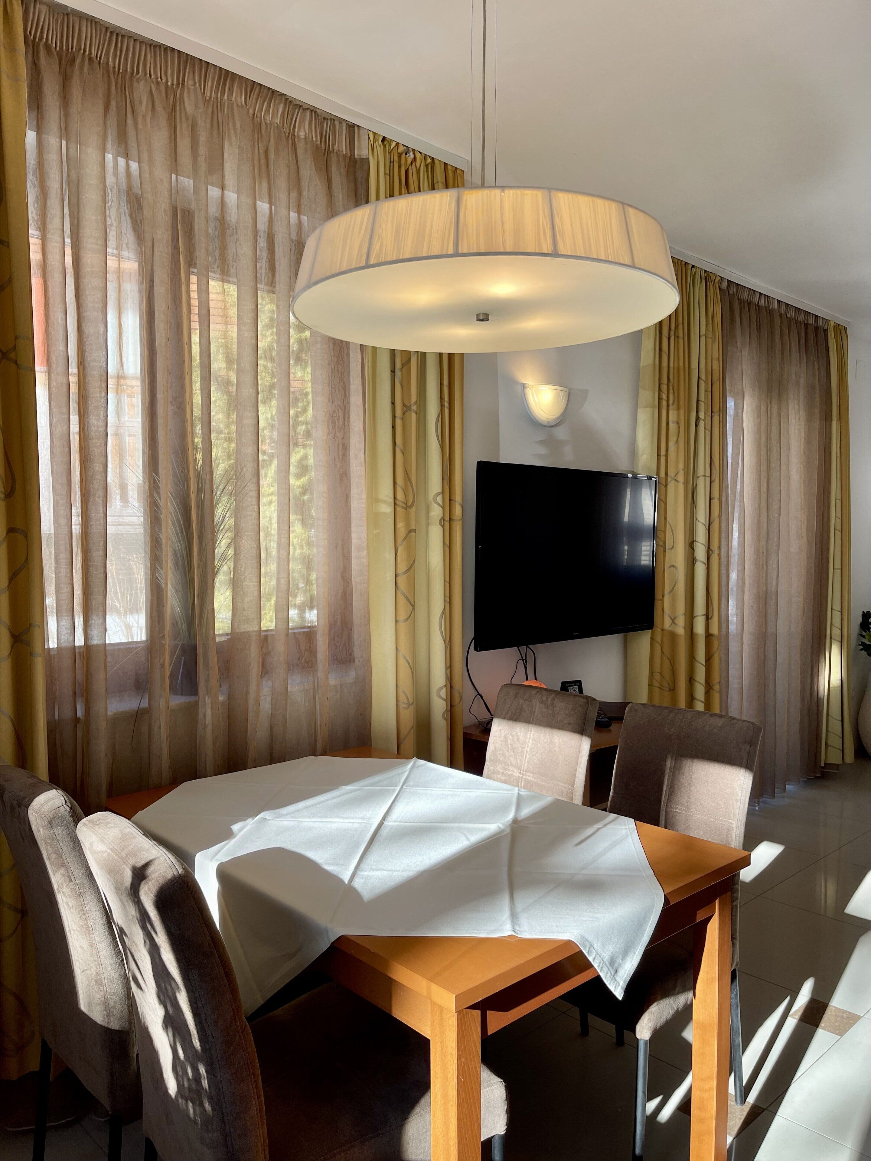 A hotel dining area with a table set for two, a television mounted on the wall, and a large ceiling light fixture.