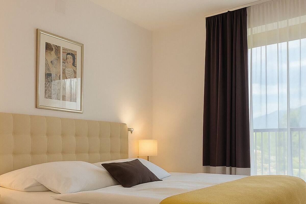 A hotel room with a large bed, a headboard, a bedside table with a lamp, a window with curtains, and a framed picture on the wall.