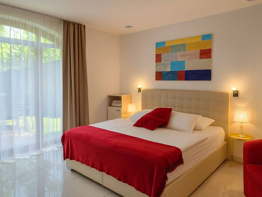 A hotel room with a large bed, two bedside tables with lamps, a colorful abstract painting on the wall, and a view of greenery through sheer curtains.