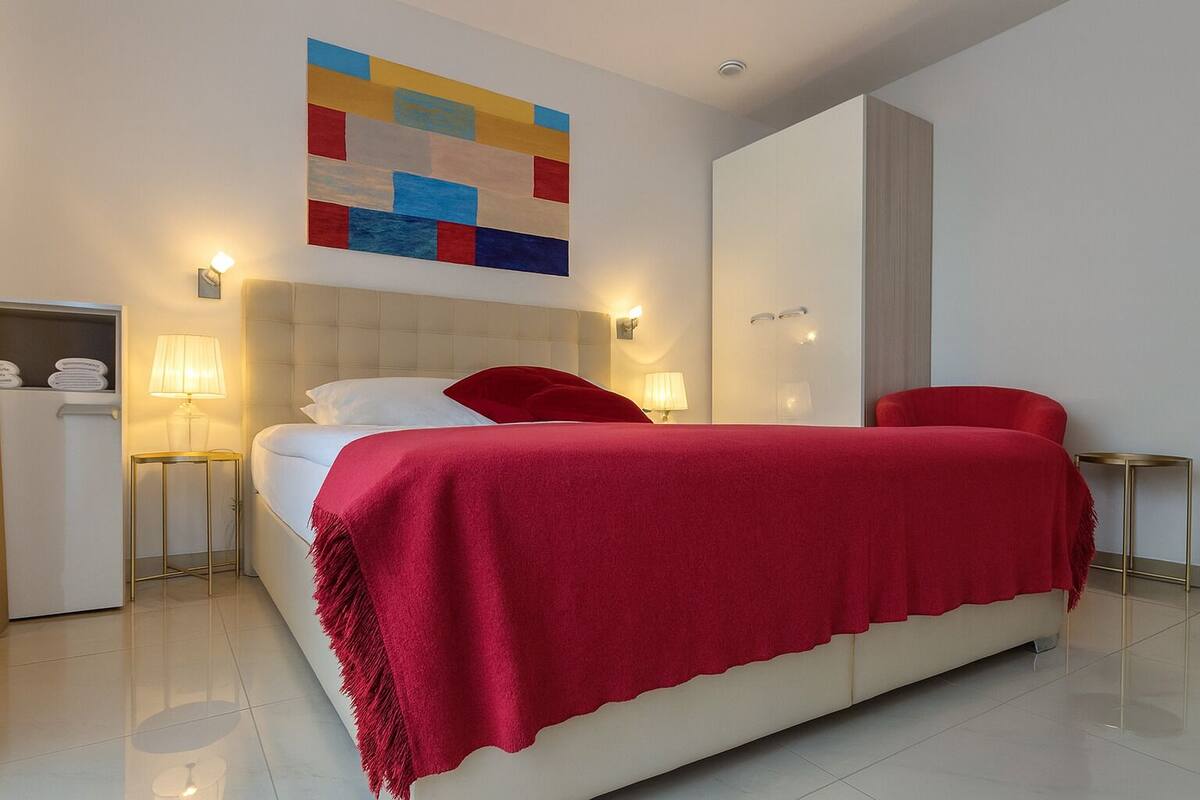 A modern hotel room with a large bed, a red throw blanket, and a colorful abstract painting above the bed.