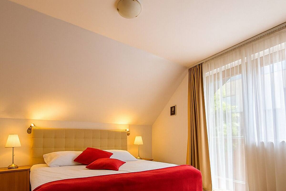 A hotel room with a large bed, two bedside lamps, a ceiling light, and a window with sheer curtains.