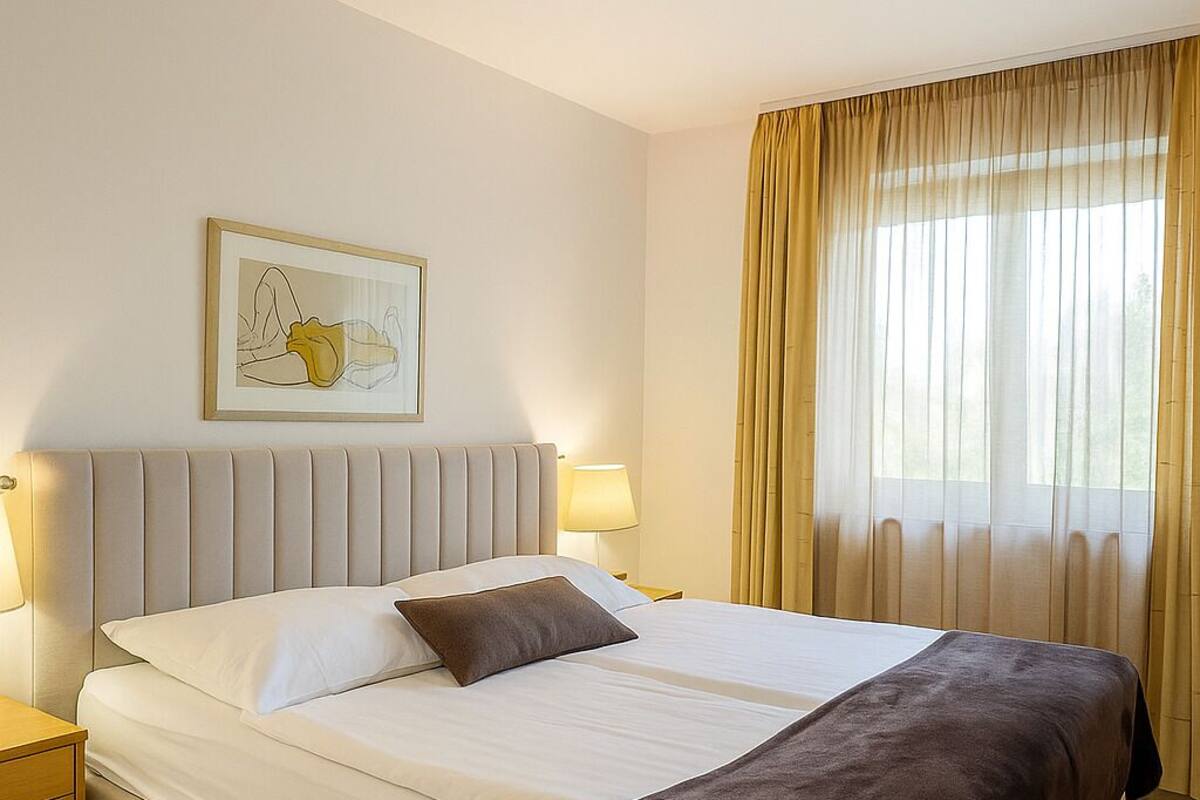 A hotel room with a large bed, a bedside table, a lamp, a framed picture on the wall, and a window with curtains.