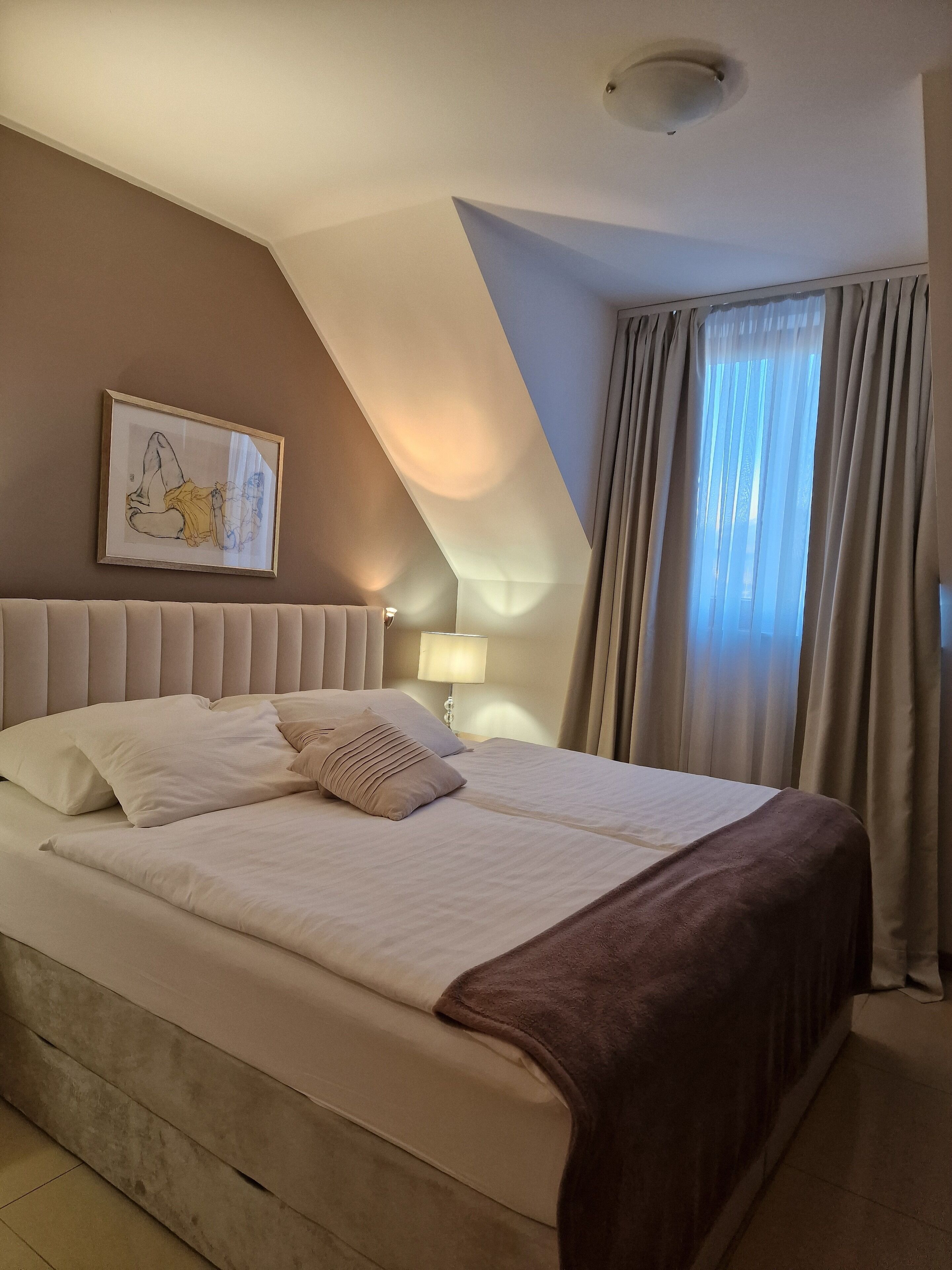 A hotel room with a large bed, a headboard, a framed artwork on the wall, a bedside lamp, and a window with curtains.
