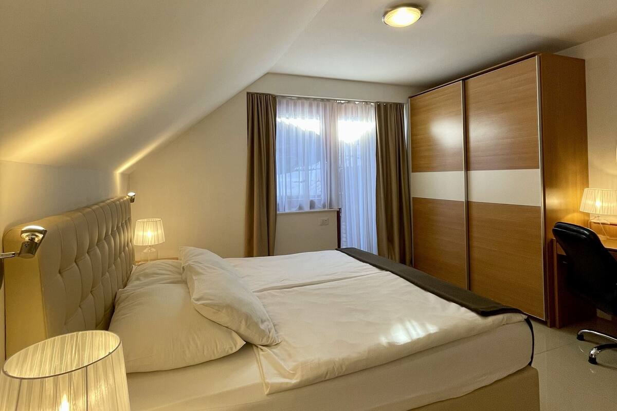 A hotel room with a large bed, a desk with a lamp, a wardrobe, and a window with curtains.