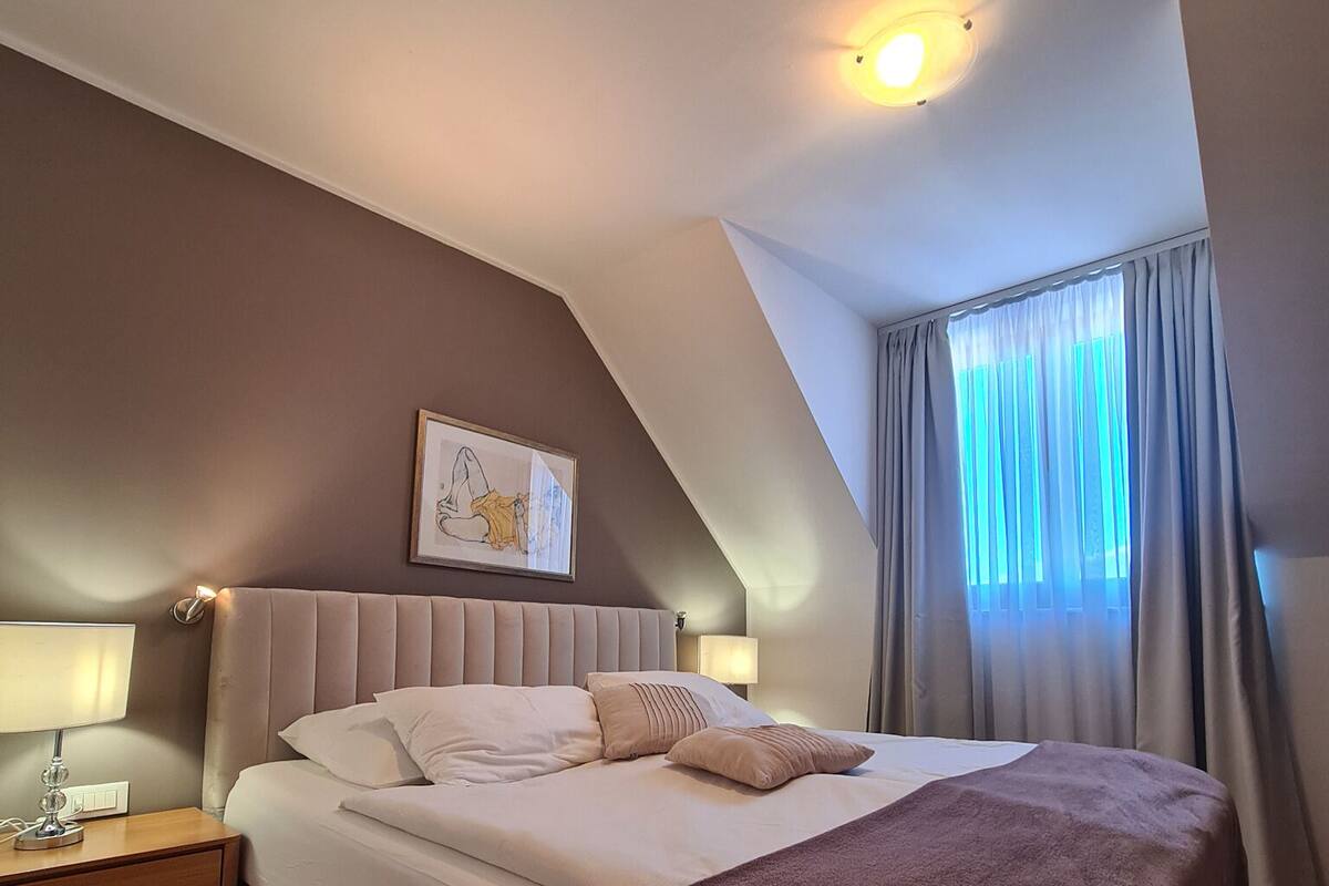 A bedroom with a bed, bedside tables, a nightstand, a framed picture on the wall, and a window with curtains.