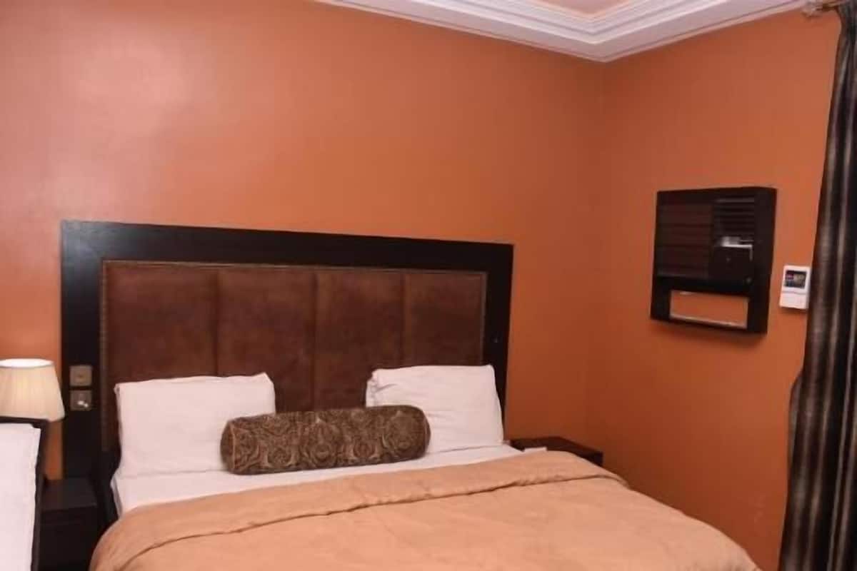 A hotel room with a large bed, a nightstand, a lamp, and a wall-mounted air conditioning unit.
