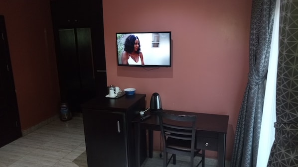A small room with a television mounted on the wall, a desk, a chair, and a kitchenette area with a coffee maker and cups. - Hard Break Hotel and Suite (Enugu)