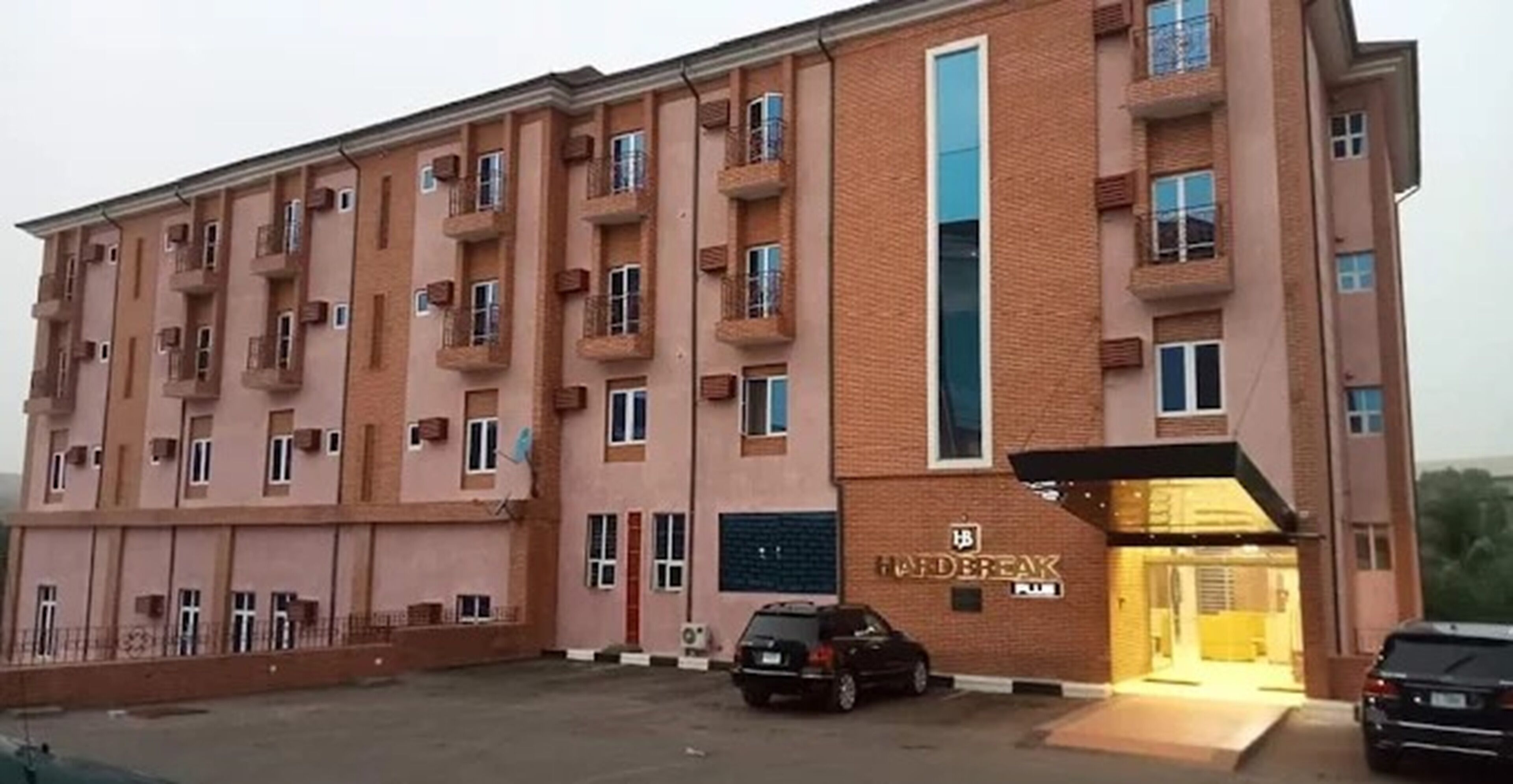 Un edificio de varios pisos con una entrada central, un letrero sobre la puerta y un estacionamiento con coches.