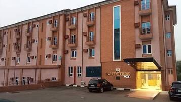 Un edificio de varios pisos con una entrada central, un letrero sobre la puerta y un estacionamiento con coches.