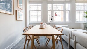 Un espace repas moderne avec une table et des chaises en bois, de grandes fenêtres équipées de stores et des œuvres d’art encadrées accrochées aux murs.