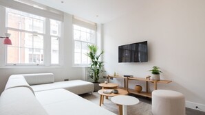 Un salon moderne avec un canapé d’angle, une table basse ronde, un téléviseur fixé au mur et une plante en pot.