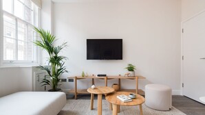 Une pièce moderne et bien éclairée, dotée d’une grande fenêtre, d’un téléviseur à écran plat, d’un bureau en bois avec deux chaises, d’une petite table ronde, d’un canapé blanc et d’une plante en pot.