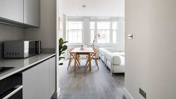 Une cuisine moderne dotée d’un four à micro-ondes encastré, un coin repas avec une table et des chaises, et un canapé blanc dans une pièce lumineuse avec de grandes fenêtres.