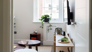 A small, well-lit room with a wooden table, a white chair, a small round table, and a potted plant on a windowsill.
