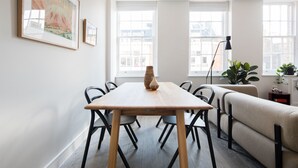 A modern living room with a wooden dining table, black chairs, a beige sofa, and a large window with blinds.