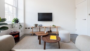 A modern living room with a large window, a flat-screen TV mounted on the wall, a wooden coffee table, and a round ottoman.