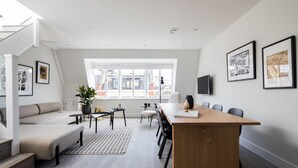 A modern living room with a sofa, a dining table, and a TV. There are framed pictures on the wall and a window with a view of buildings. - Spacious Leicester Sq 3BR Penthouse (London)