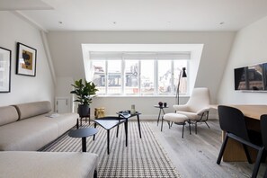 A modern living room with a sofa, a dining table, a chair, and a large window with a city view. - Spacious Leicester Sq 3BR Penthouse (London)