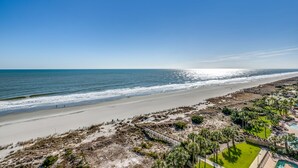 A beach view with a clear sky, a sandy beach, and a resort area with a pool and lounge chairs. - 🌊🌞Lazy River + Indoor/Outdoor Pools | Oceanfront | South Wind 1101 (Myrtle Beach)
