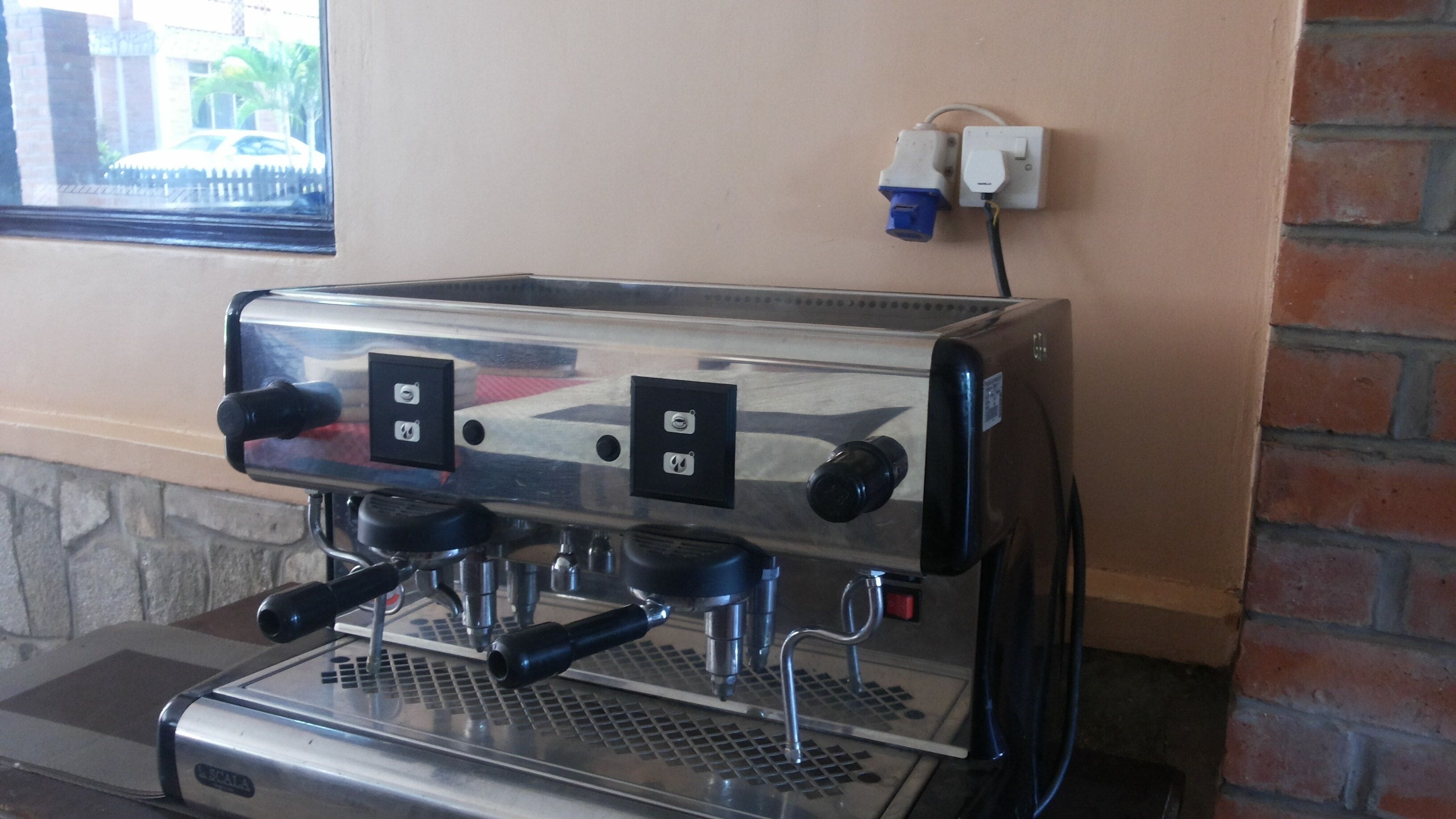 An espresso machine with two control knobs and a digital display, connected to a power outlet on a brick wall.