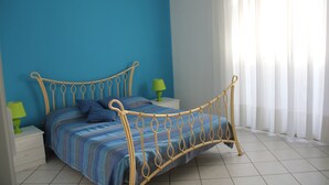 A bed with a metal frame, a blue-striped bedspread, and pillows, a green lamp on a bedside table, and a window with sheer curtains. - Apartment le Maschere (Marina di Modica)