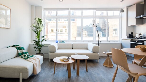 Un salon moderne comprenant un canapé, deux petites tables rondes, une table à manger avec des chaises, une grande fenêtre donnant sur des immeubles et un coin cuisine avec une cuisinière et des placards.