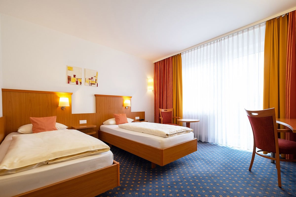 Hotellihuone, jossa on kaksi sänkyä, työpöytä, tuoli ja suuri ikkuna, jossa on verhot.
