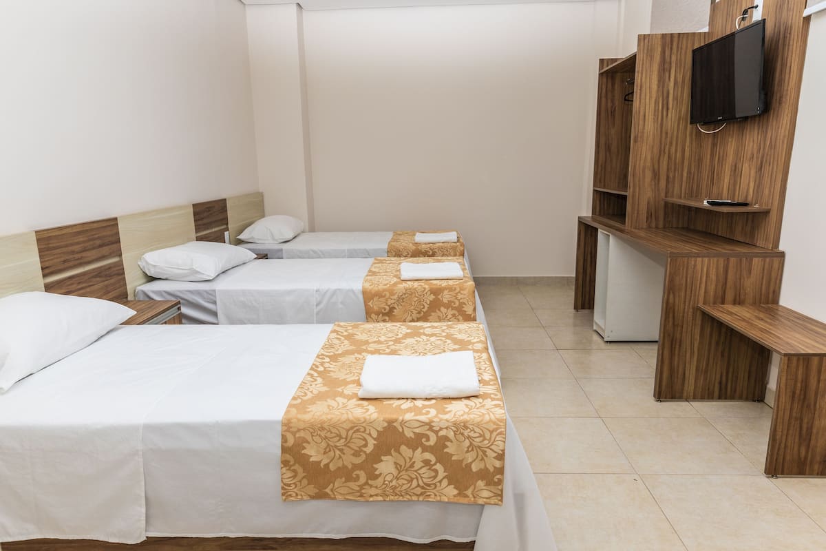 Habitación de hotel con tres camas individuales, un televisor montado en un mueble de madera y suelo de baldosas blancas.
