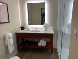 Un baño moderno con lavamanos blanco, espejo y tocador de madera. Hay toallas y artículos de aseo en el tocador, y un secador de pelo en el suelo.