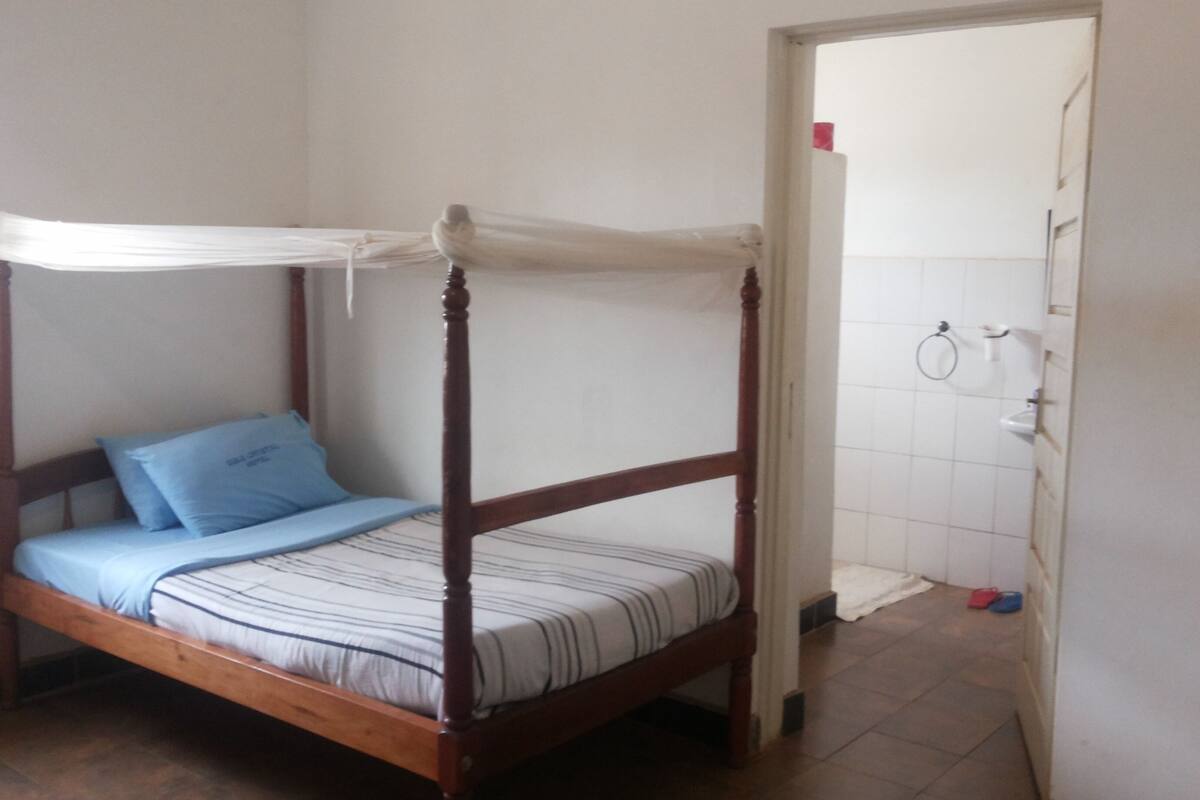 A single bed with a wooden frame and a canopy, a blue pillow, and a striped bedspread. There is a small bathroom with a toilet, a towel rack, and a shower area.